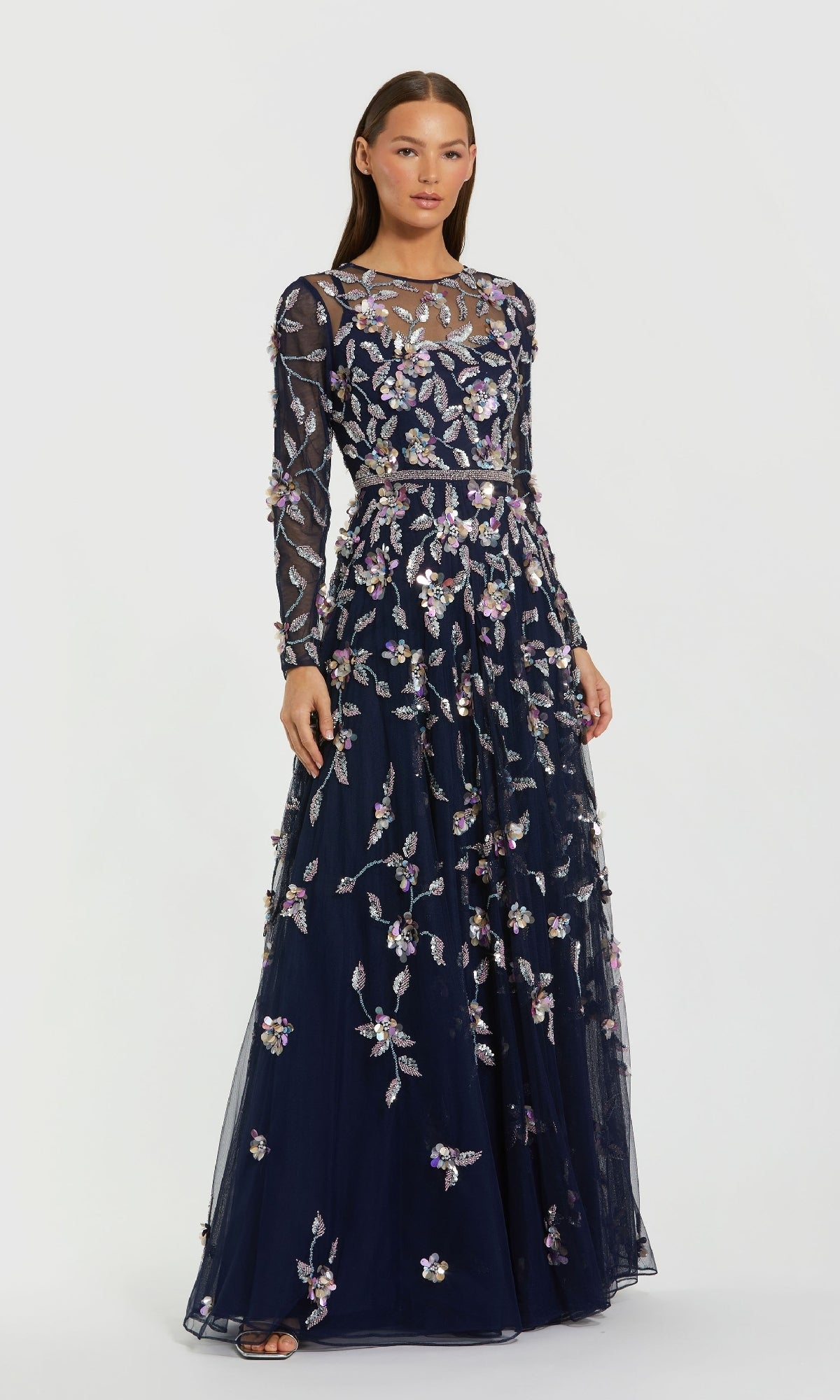 A woman models the Mac Duggal 6183 Long Sleeve Beaded Formal Gown, featuring navy blue fabric, sheer long sleeves, and floral embroidery, standing against a plain white background.